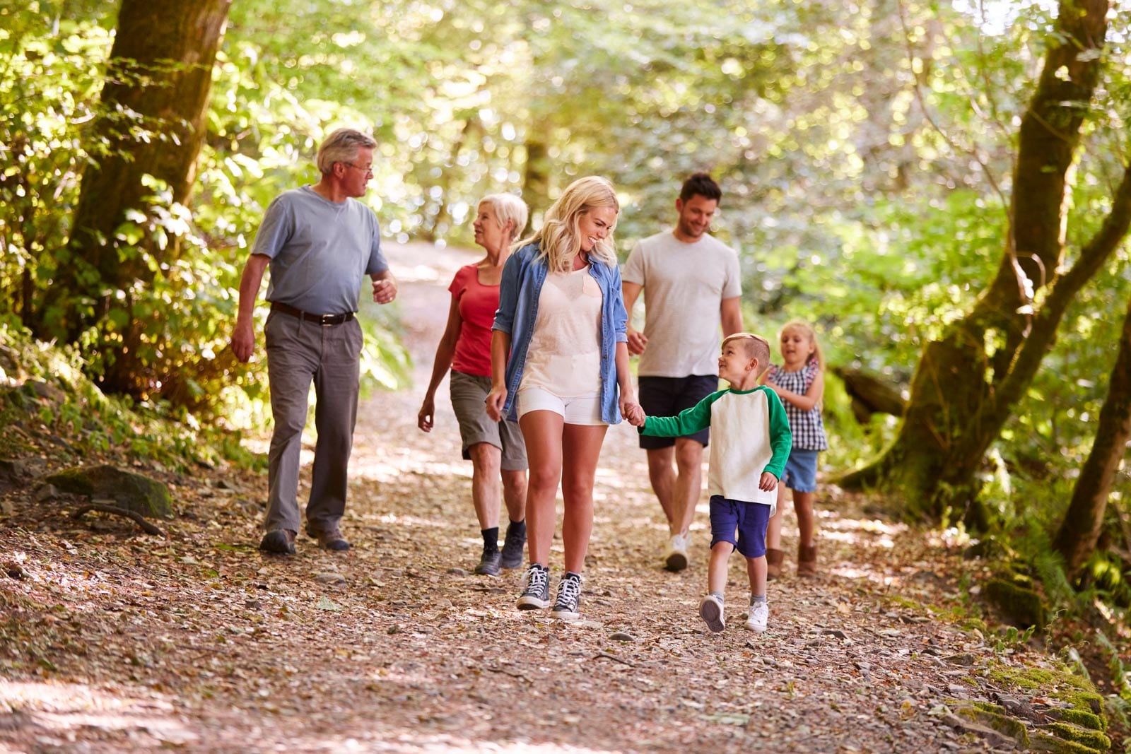 Three adults and three children walk together on a wooded trail, surrounded by trees and sunlight filtering through the leaves.