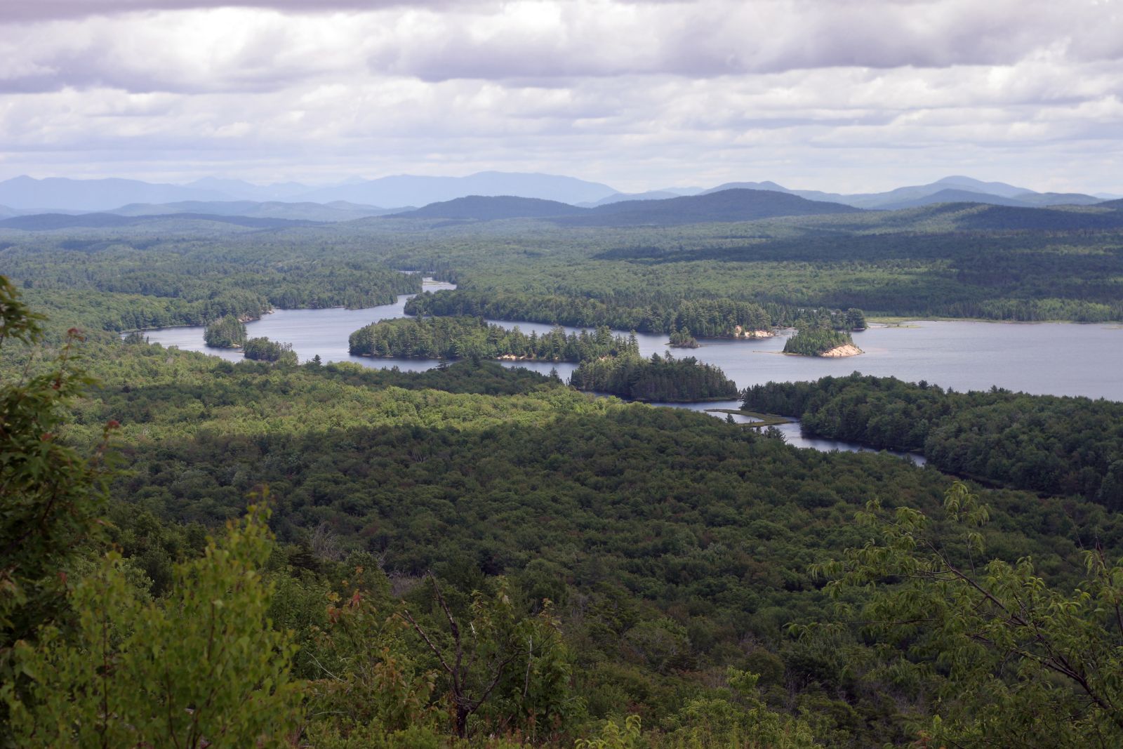 Scenic view of a forested landscape with a large lake, multiple islands, and distant mountains under a cloudy sky.