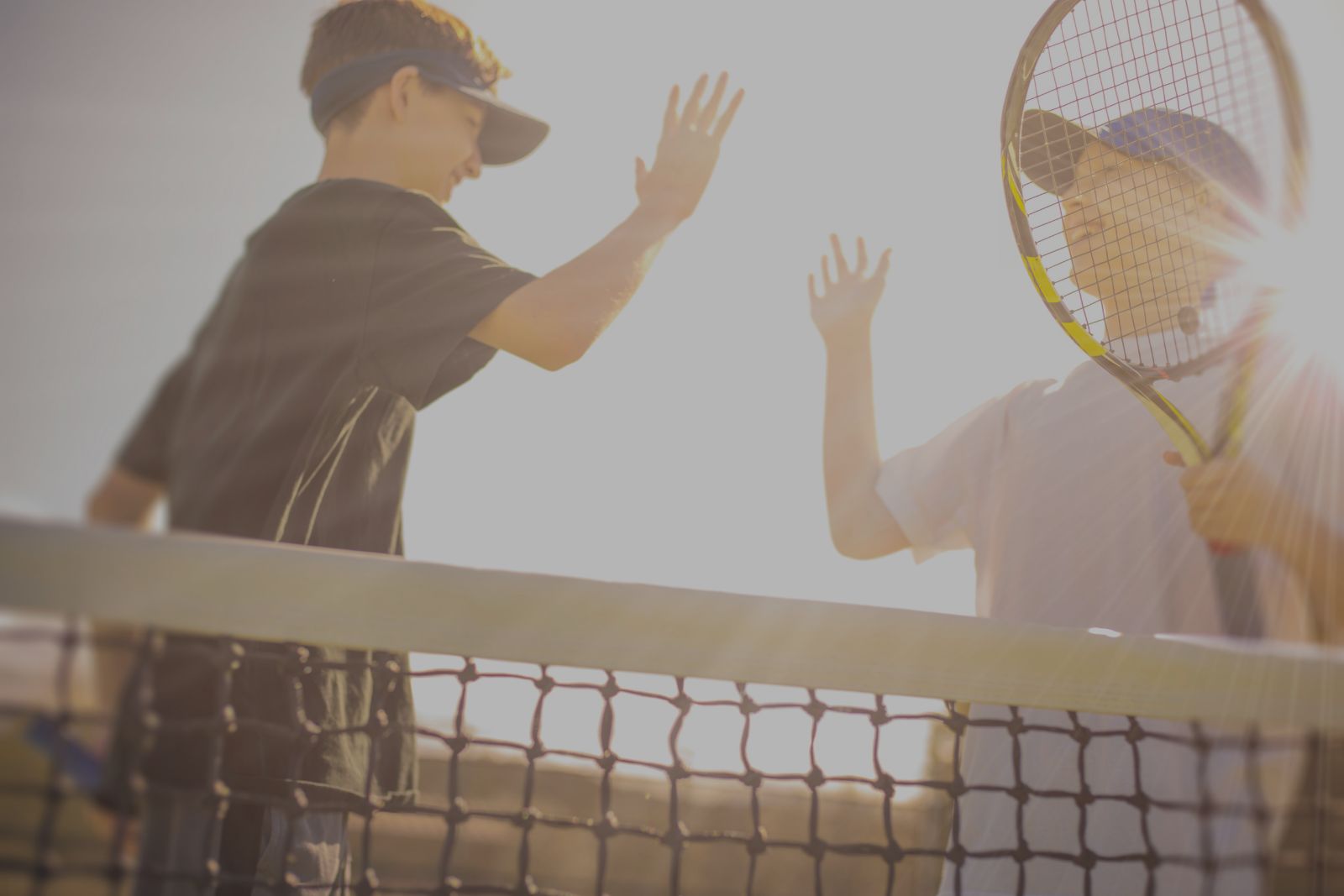 Two boys wearing visors and sports clothes share a high-five over a tennis net, one holding a racket, with sunlight shining behind them.