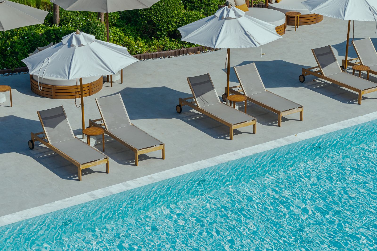 Several empty lounge chairs with white umbrellas are lined up beside a clear blue swimming pool on a sunny day.