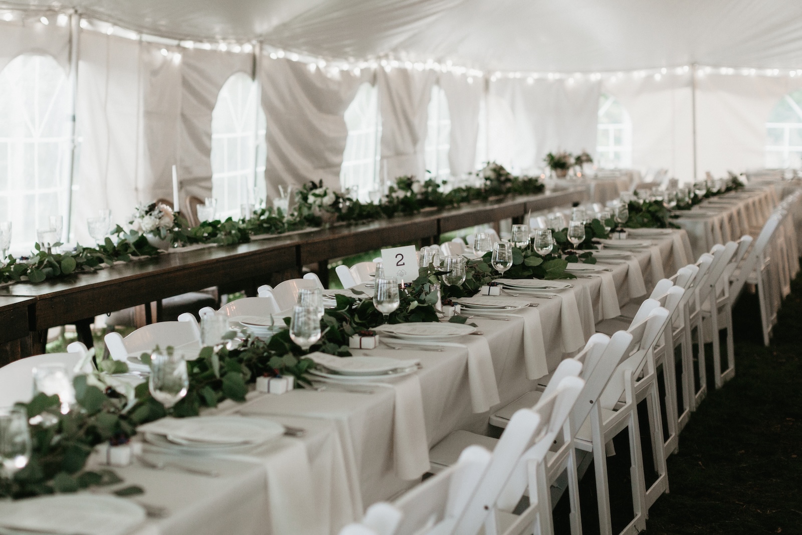 Long tables set for an event under a white tent, with white tablecloths, green garlands, plates, glassware, and a table number sign visible.