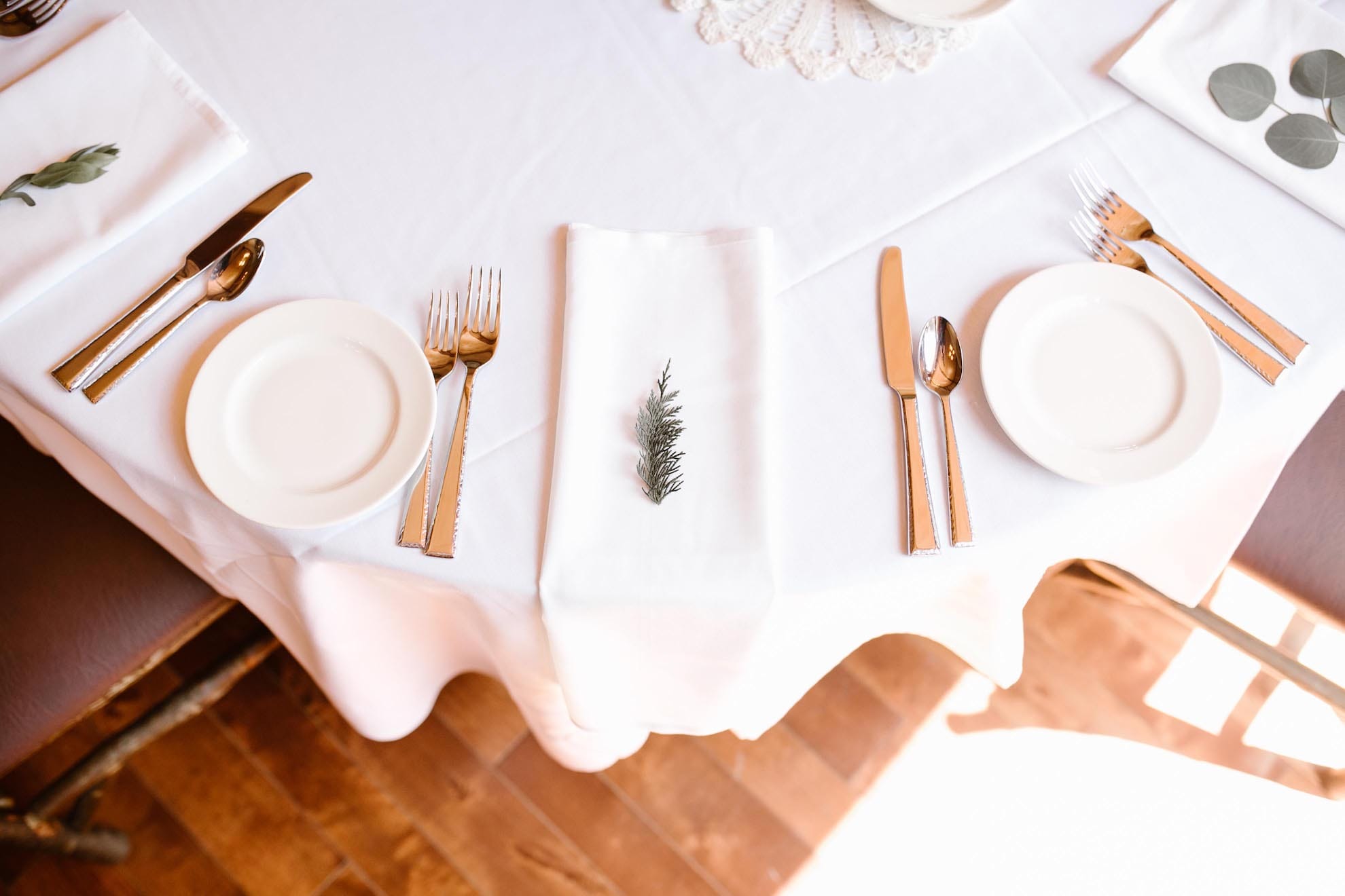 A neatly set dining table with white plates, silver cutlery, white napkins, and a small sprig of greenery on one napkin, all on a white tablecloth.