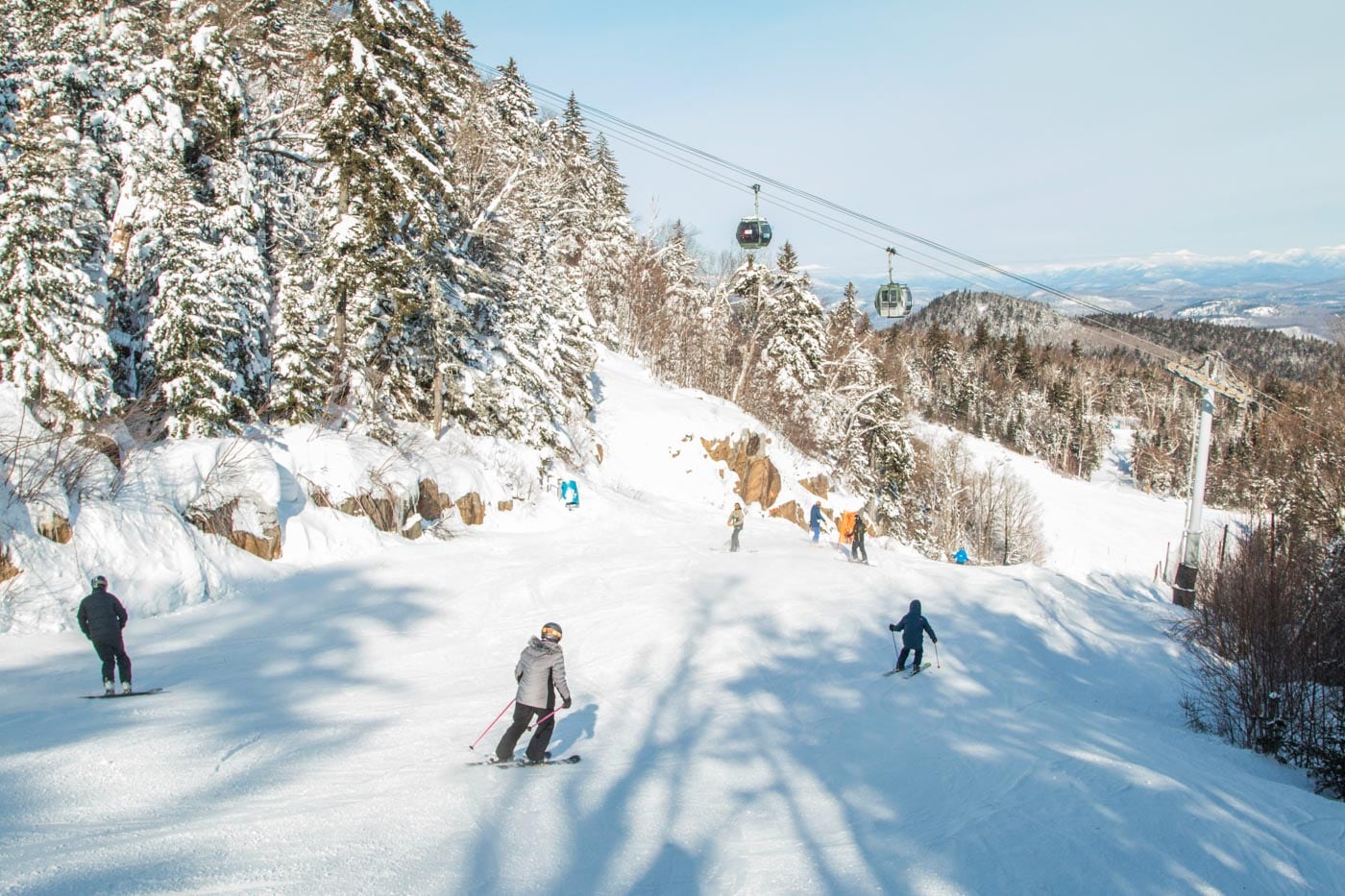 People skiing down a snowy mountain slope surrounded by trees, with a ski lift and gondolas overhead on a clear day.