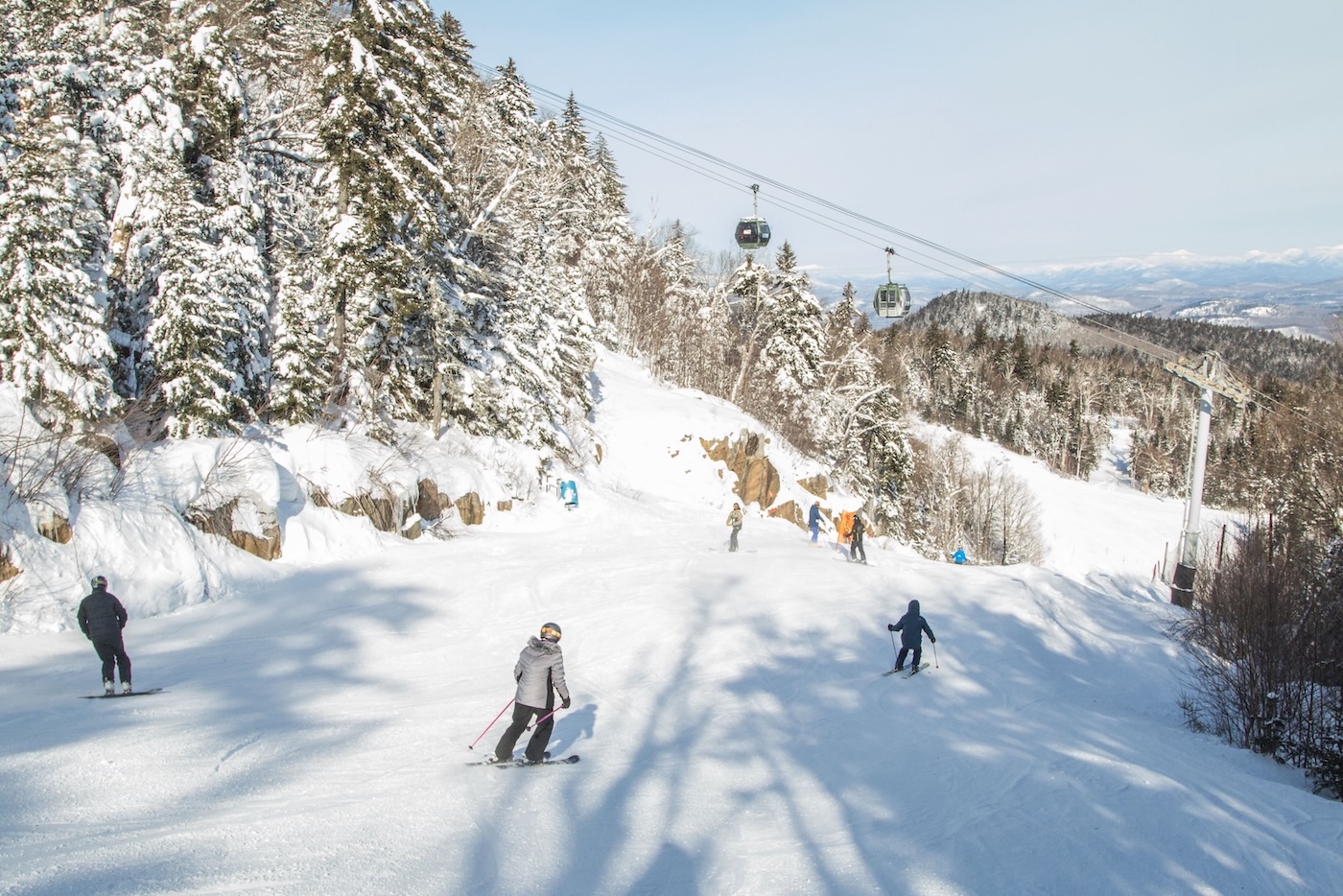 People skiing down a snowy slope surrounded by trees, with a ski lift and gondolas overhead under a clear sky.
