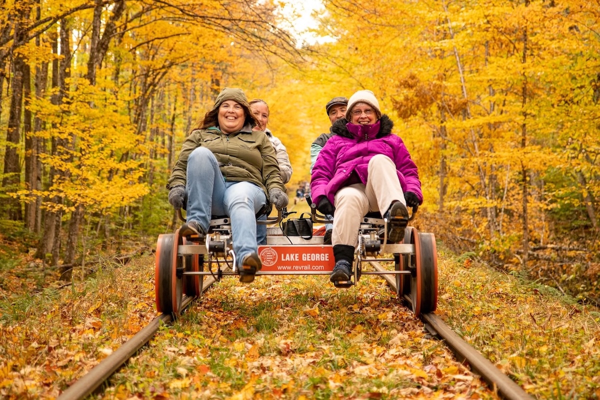 Four people pedal a rail bike along tracks through a forest with vibrant yellow autumn foliage.