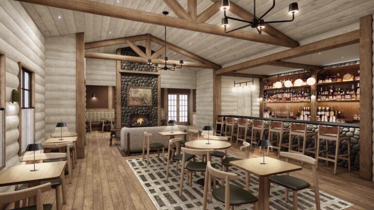 Spacious lodge-style restaurant with wooden beams, stone fireplace, modern bar, and neatly arranged tables and chairs on a patterned rug.