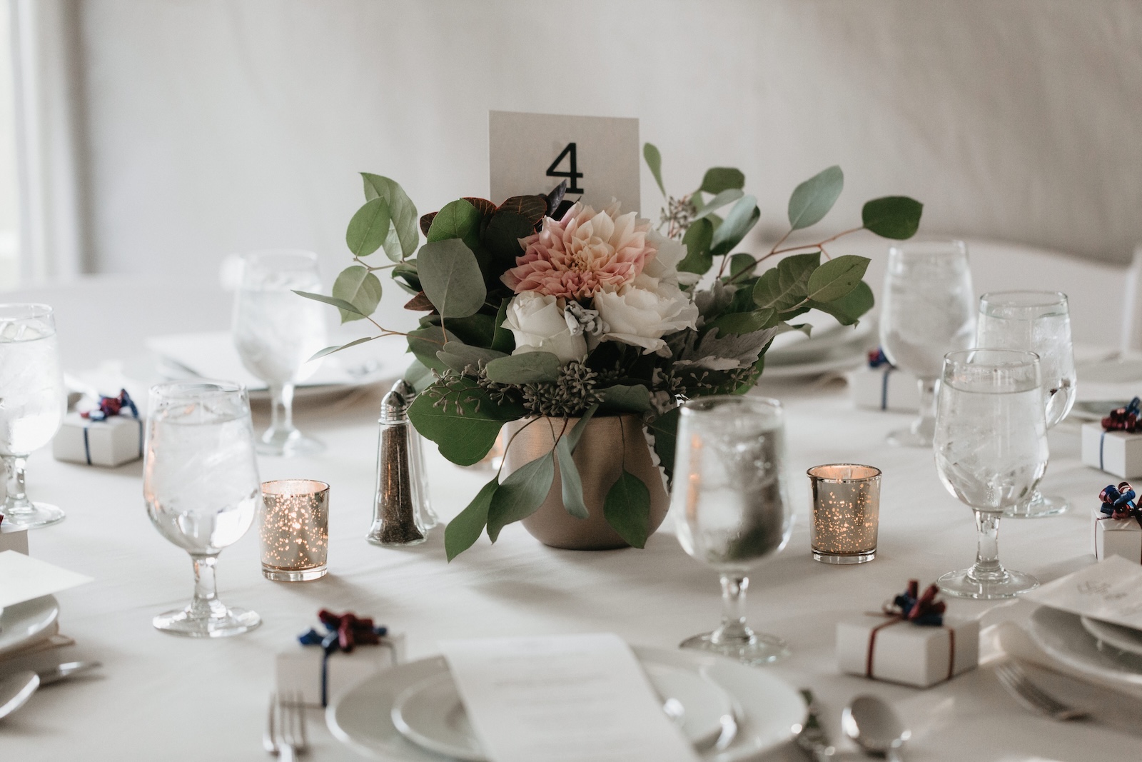 A table set for a formal event with a floral centerpiece, candles, wrapped gifts, water glasses, and a table number card displaying the number 4.