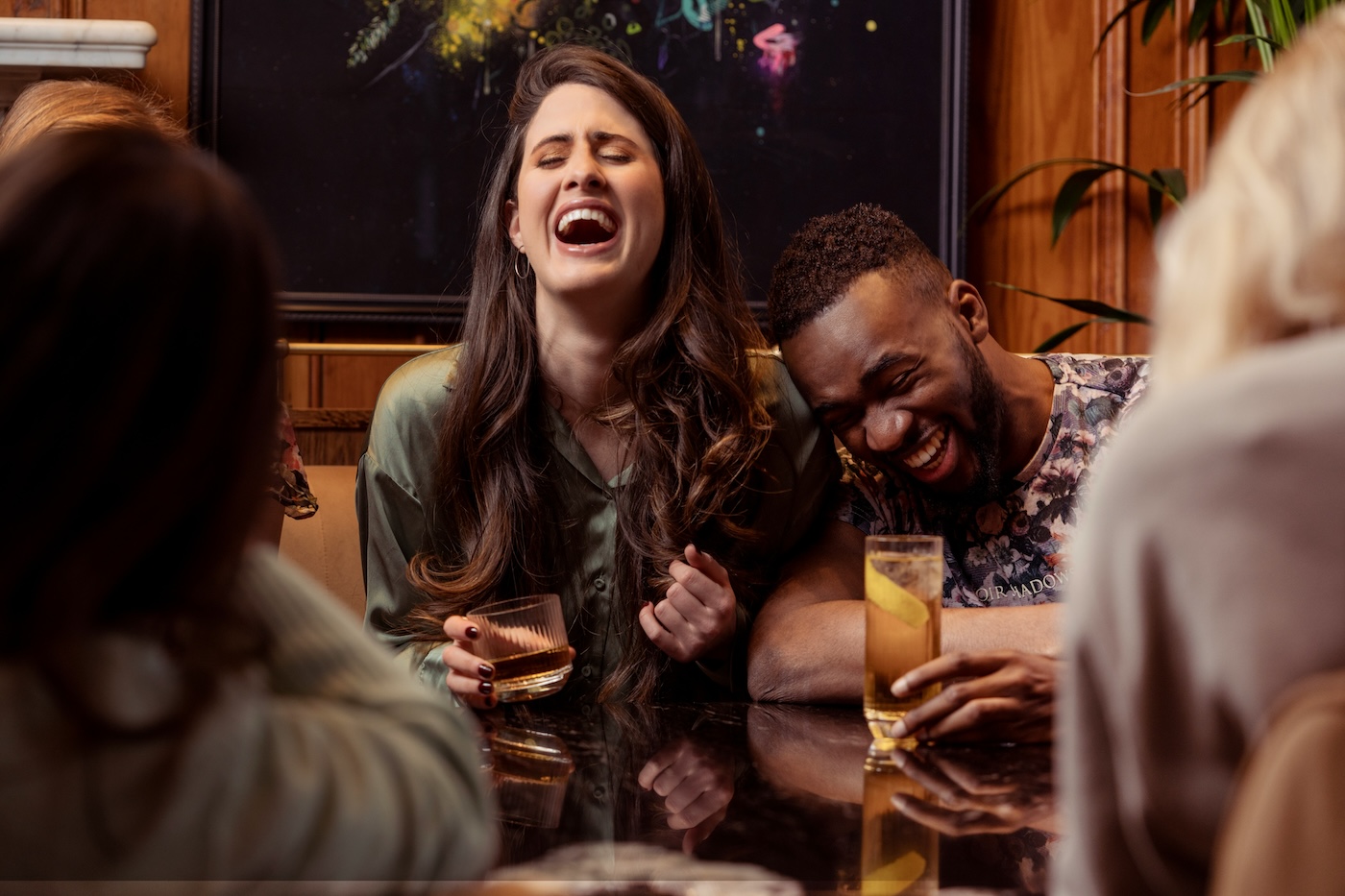 Two people sit at a table laughing while holding drinks, surrounded by others in a lively indoor setting.