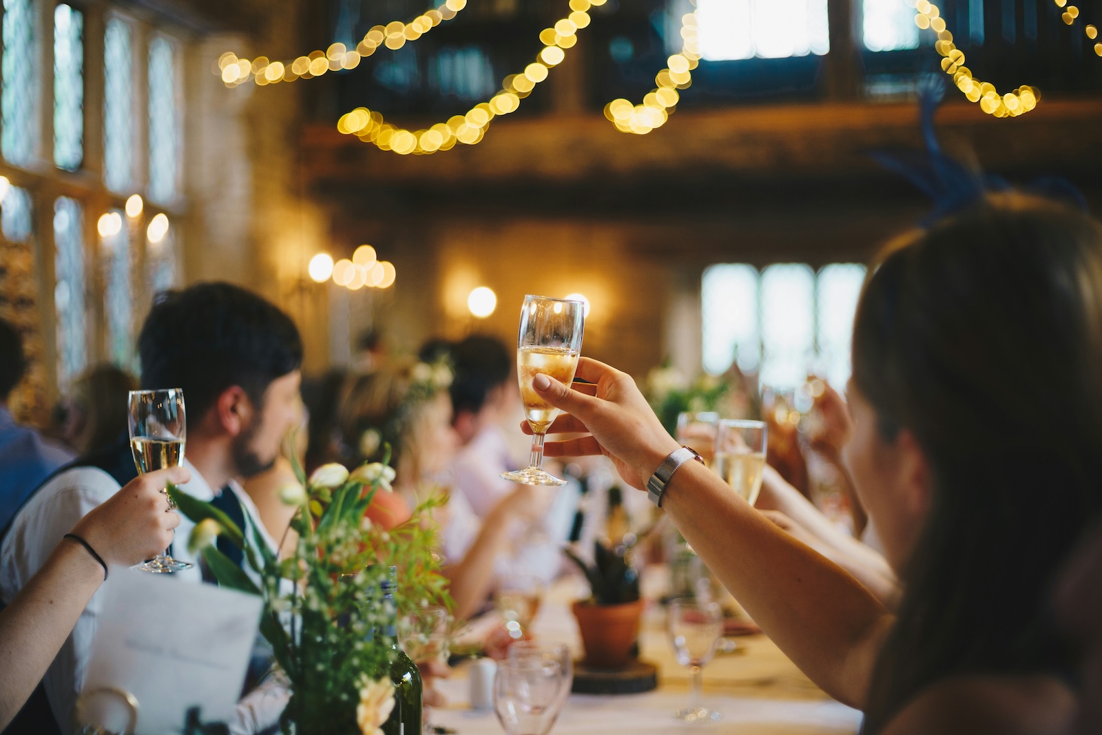 People seated at a decorated table raise glasses in a toast, with warm lighting and string lights in the background.